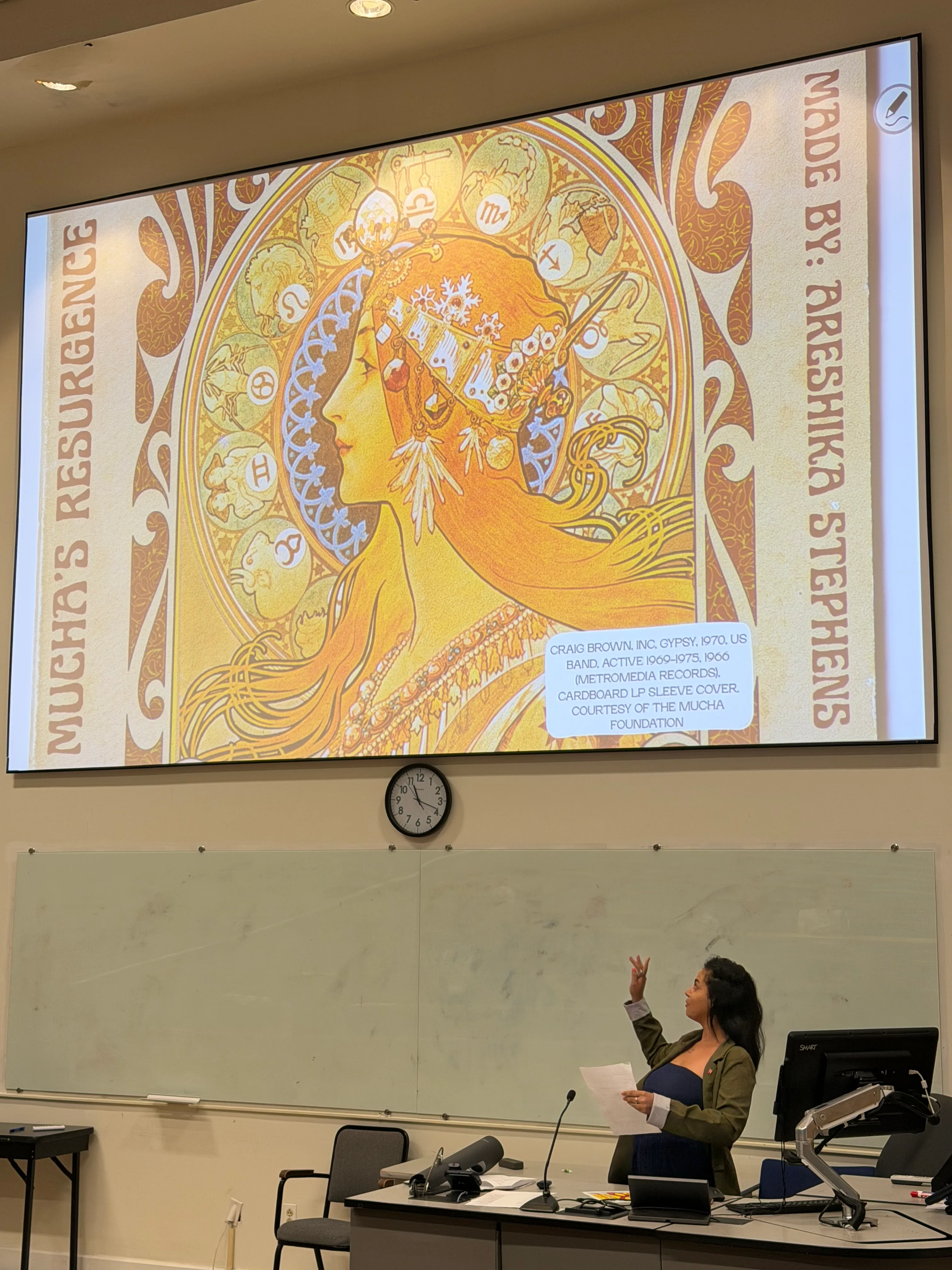 Areshika presenting a lecture with a projected Art Nouveau Mucha-style illustration titled Mucha's Resurgence